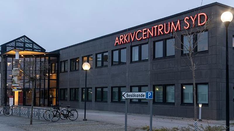 Arkivcentrum Syds moderna byggnad i Lund under kvällstid, med en fasad i mörkgrått och stora fönster. En skylt med röd text lyser upp på taket, och cykelställ samt en parkeringsskylt för besökare syns i förgrunden.